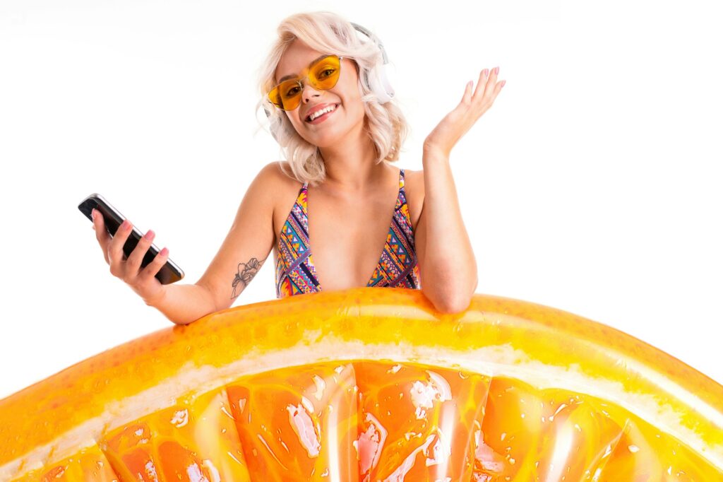 Pretty blonde caucasian female stands in swimsuit with rubber beach orange mattress, phone and